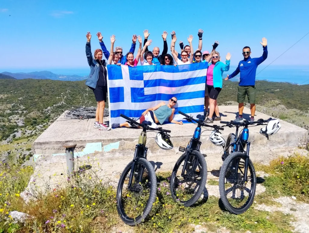 E-bike group with raised arms and a Greek flag on a hilltop above the sea